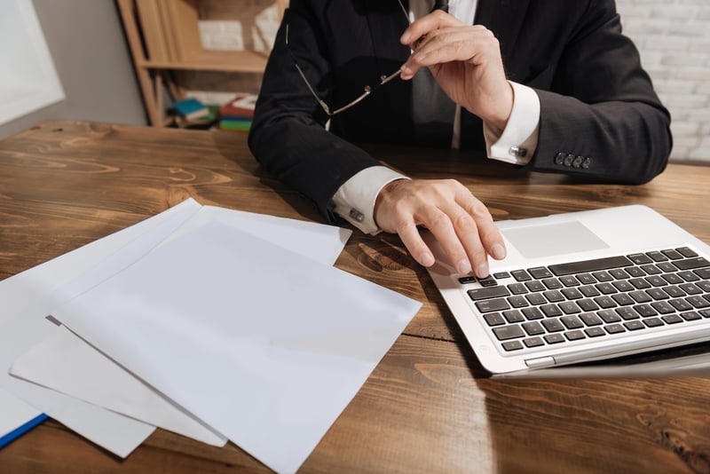 A well-dressed older worker, likely a high-income earner, sits at a wooden desk with a stack of white papers to their left and an open silver laptop on the right, actively engaged in work. The person's hands are prominent, one resting on the laptop's trackpad as if scrolling, and the other holding a pair of eyeglasses, suggesting deep thought or a break from screen work. This image contextually illustrates the article's focus on changes to 401(k) catch-up contributions for older workers earning over $145,000, particularly the shift to Roth contributions for high earners starting in 2026. The scene evokes the financial planning and adjustments these individuals may face regarding their retirement savings.