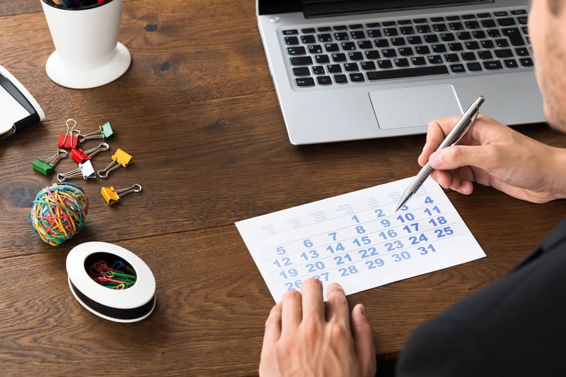 An overhead shot captures a person's hands holding a silver pen over a printed calendar page, likely marking or planning for the upcoming July 1, 2026 deadline. The calendar, displaying the days of a month, rests on a warm, dark wooden desk. To the right, a silver laptop with a visible keyboard and touchpad is open, suggesting a work or research environment. Various office supplies are scattered around the desk, including a white pen holder with a dark pen, a small pile of colorful binder clips, a multi-colored rubber band ball, and a white oval dish filled with paper clips. This detailed scene visually represents the meticulous planning and organization required as individuals and families prepare for the significant federal policy changes impacting student loan caps and repayment options beginning in 2026. Source: The College Investor