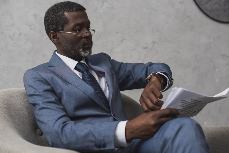 Waiting on a report A distinguished Black man in a tailored blue suit and white shirt, accented with a dark patterned tie, sits in a modern gray armchair against a textured concrete wall. He wears sophisticated gold-rimmed glasses and has neatly groomed gray hair and a beard. His expression is one of focused anticipation as he holds a stack of papers, seemingly reports or applications, in his hands, looking down. This image effectively illustrates the article's theme of "Waiting on a report" regarding the Public Service Loan Forgiveness (PSLF) Buyback backlog, reflecting the ongoing delays and the pause in monthly status reports due to the government shutdown, leaving over 74,000 pending applications in limbo for borrowers awaiting updates on their student loans.