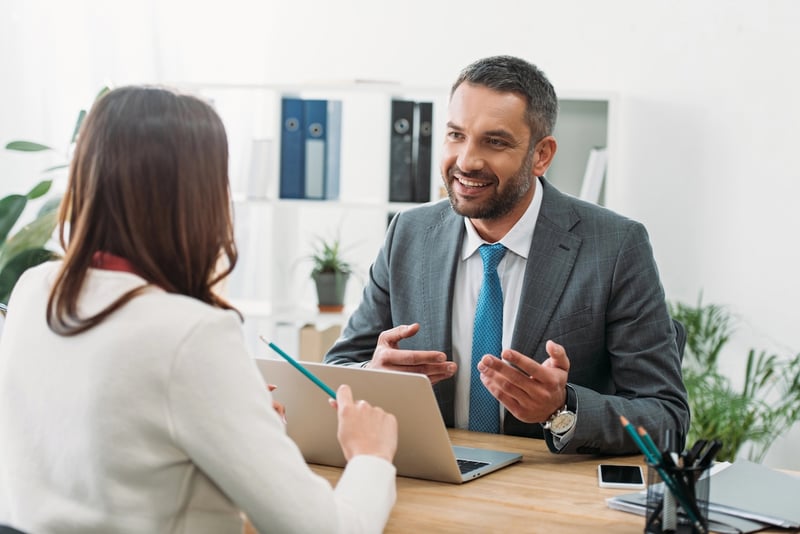 Meeting With A Student Loan Professional A smiling businessman in a grey suit and blue tie gestures while speaking to a client in a bright office. This image represents the role of financial professionals—specifically Certified Financial Planners (CFPs) or student loan consultants—who provide strategic advice to borrowers navigating complex repayment options, forgiveness plans, and long-term financial goals. Source: The College Investor