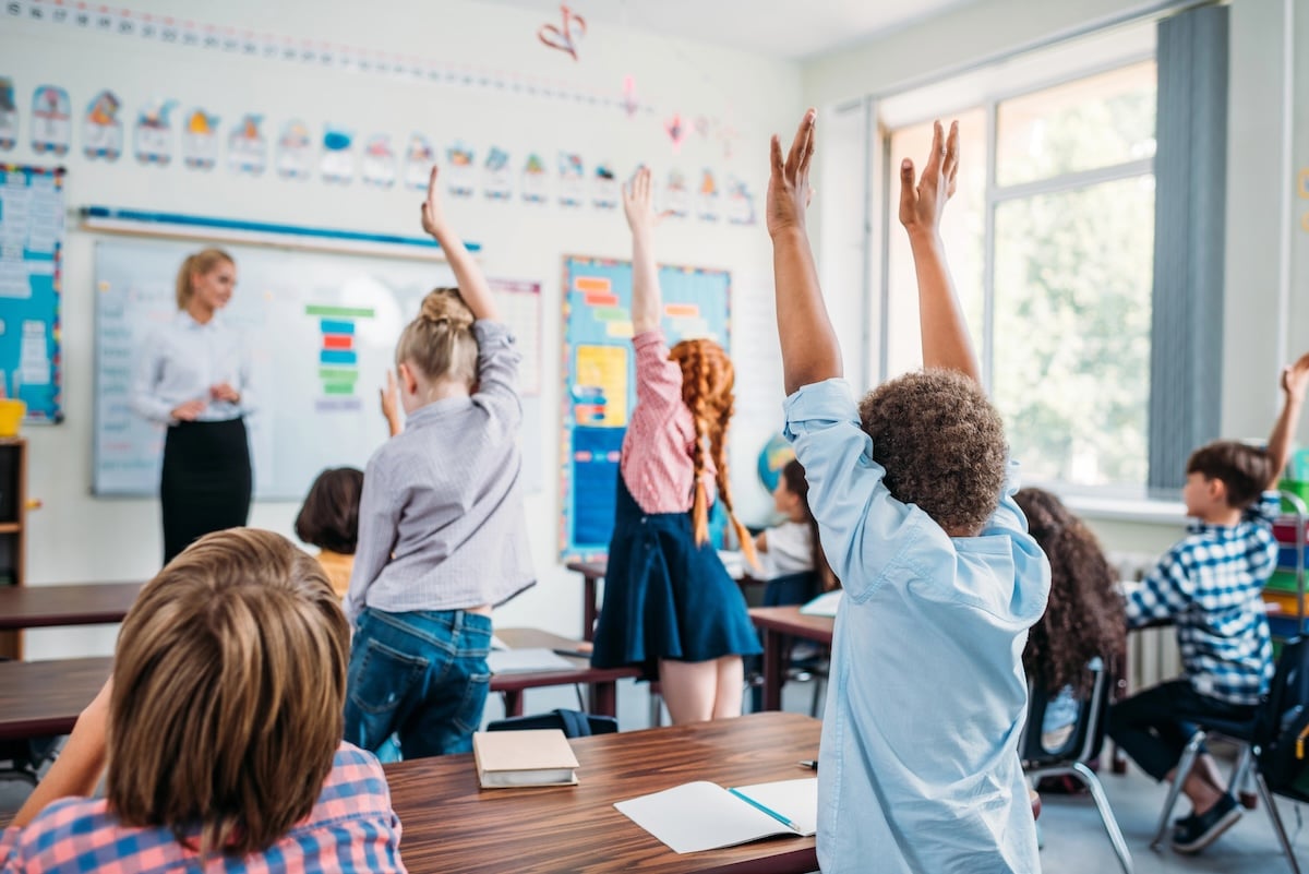 Elementary school students raising their hands in a classroom as a teacher stands at the whiteboard